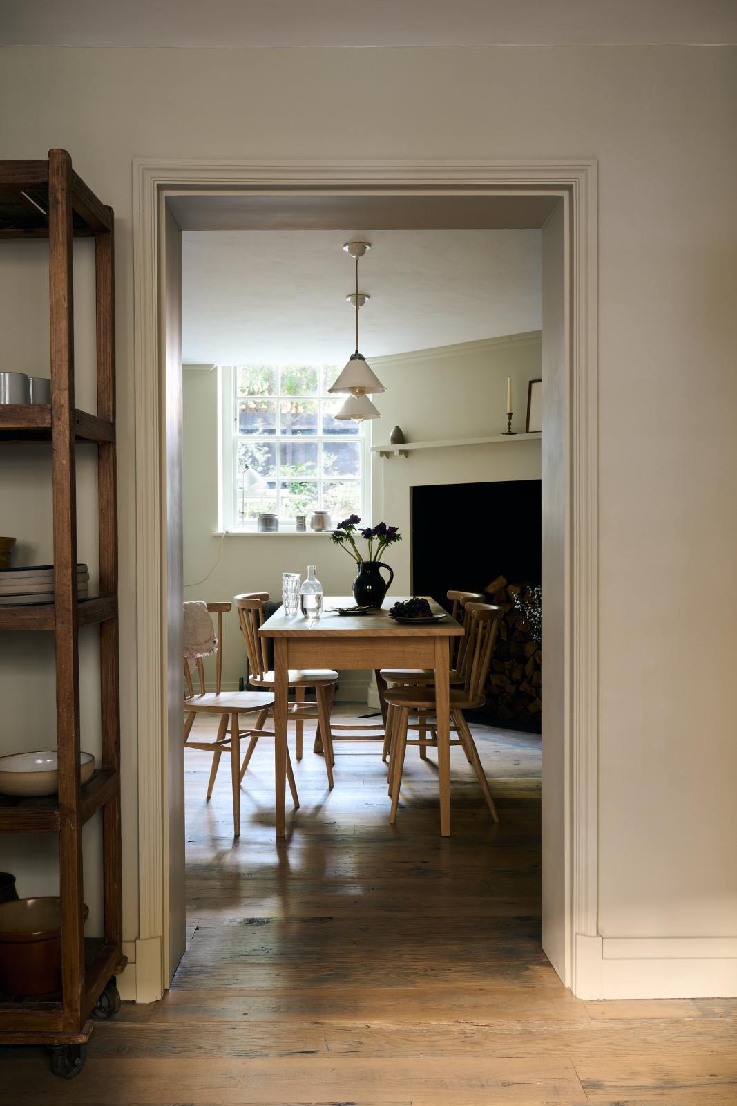 Looking through the archway in the kitchen, beside a large wooden shelf displaying a thoughtful selection of muted ceramics and mugs, the dining room comes into view with a pale wooden dining table and matching chairs.