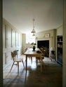 Looking through the archway in the kitchen, beside a large wooden shelf displaying a thoughtful selection of muted ceramics and mugs, the dining room comes into view with a pale wooden dining table and matching chairs. A large open fireplace adds warmth to the space, accompanied by a generous storage cupboard and cabinets. A freestanding open larder showcases beautifully simple cream, green, and reddish-brown ceramic vases and jugs.