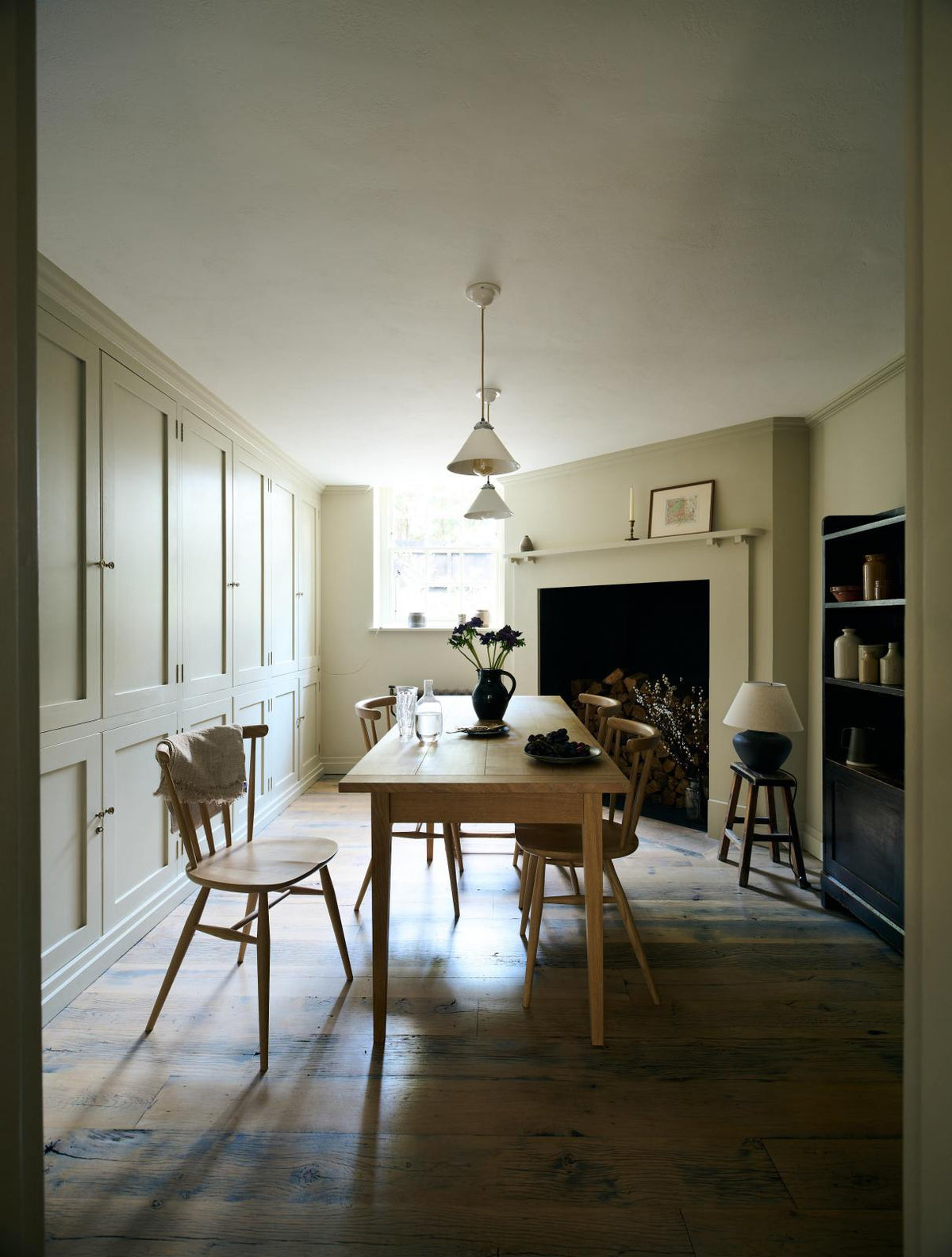Looking through the archway in the kitchen, beside a large wooden shelf displaying a thoughtful selection of muted ceramics and mugs, the dining room comes into view with a pale wooden dining table and matching chairs. A large open fireplace adds warmth to the space, accompanied by a generous storage cupboard and cabinets. A freestanding open larder showcases beautifully simple cream, green, and reddish-brown ceramic vases and jugs.
