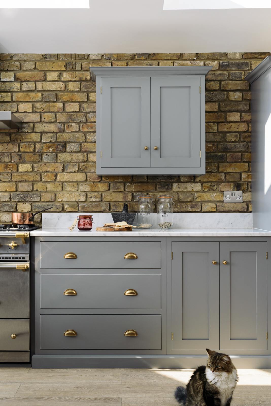 The edge of a soft industrial grey Shaker kitchen features an L-shaped sink and cooker run, with a Carrara marble worktop, wall cupboards and a fluffy kitty. Adorned with a weathered brick wall installation, copper pendant lights, dried ingredients in glass jars, jam, and a stone pestle and mortar, it creates a modern and functional kitchen for enthusiastic food connoisseurs.