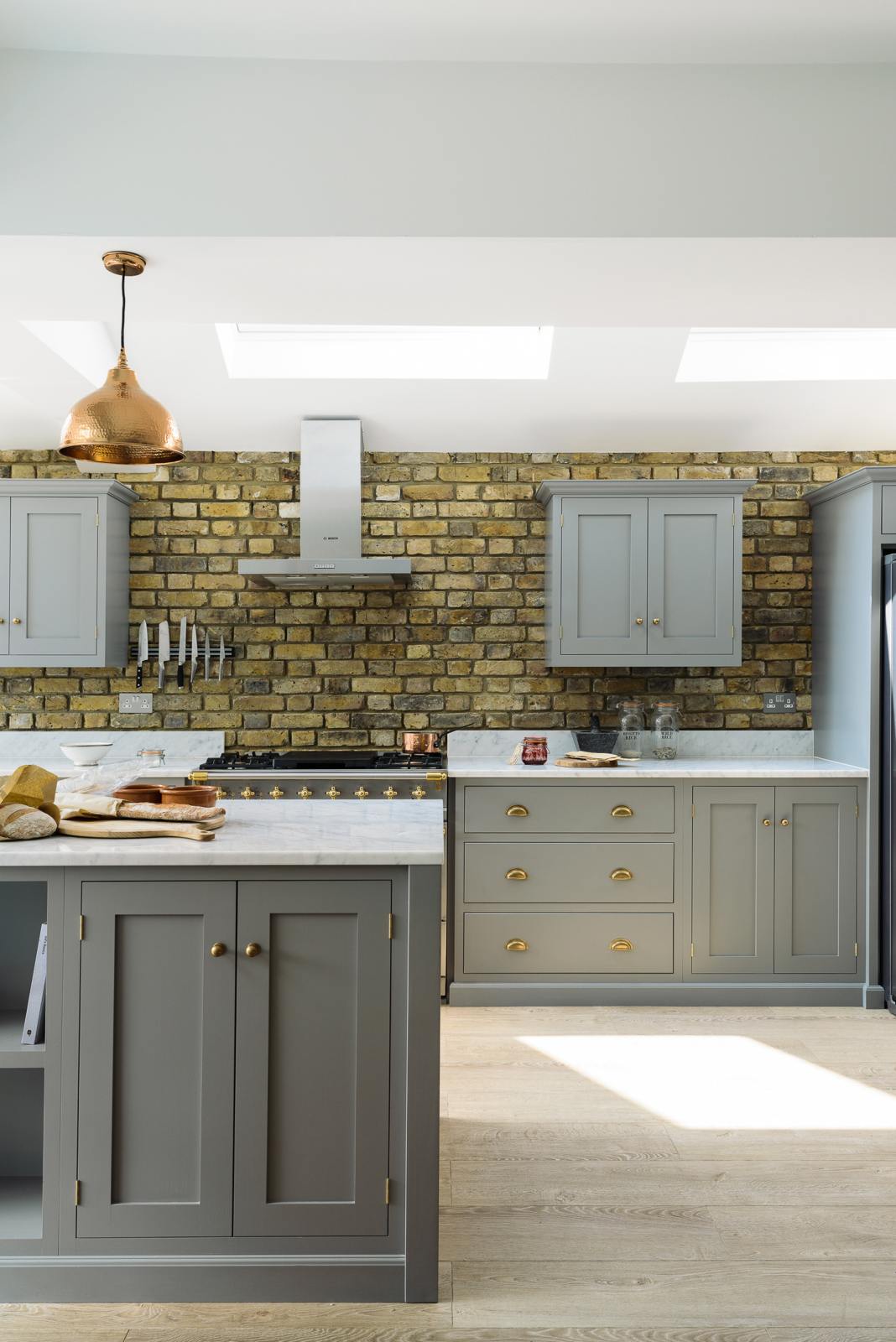 The edge of a soft industrial grey Shaker kitchen features an L-shaped sink and cooker run with a Carrara marble worktop, a kitchen island, and wall cupboards. Adorned with a weathered brick wall installation, copper pendant lights, simple ceramics, and fresh baguettes, it creates a modern and functional kitchen for enthusiastic food connoisseurs.