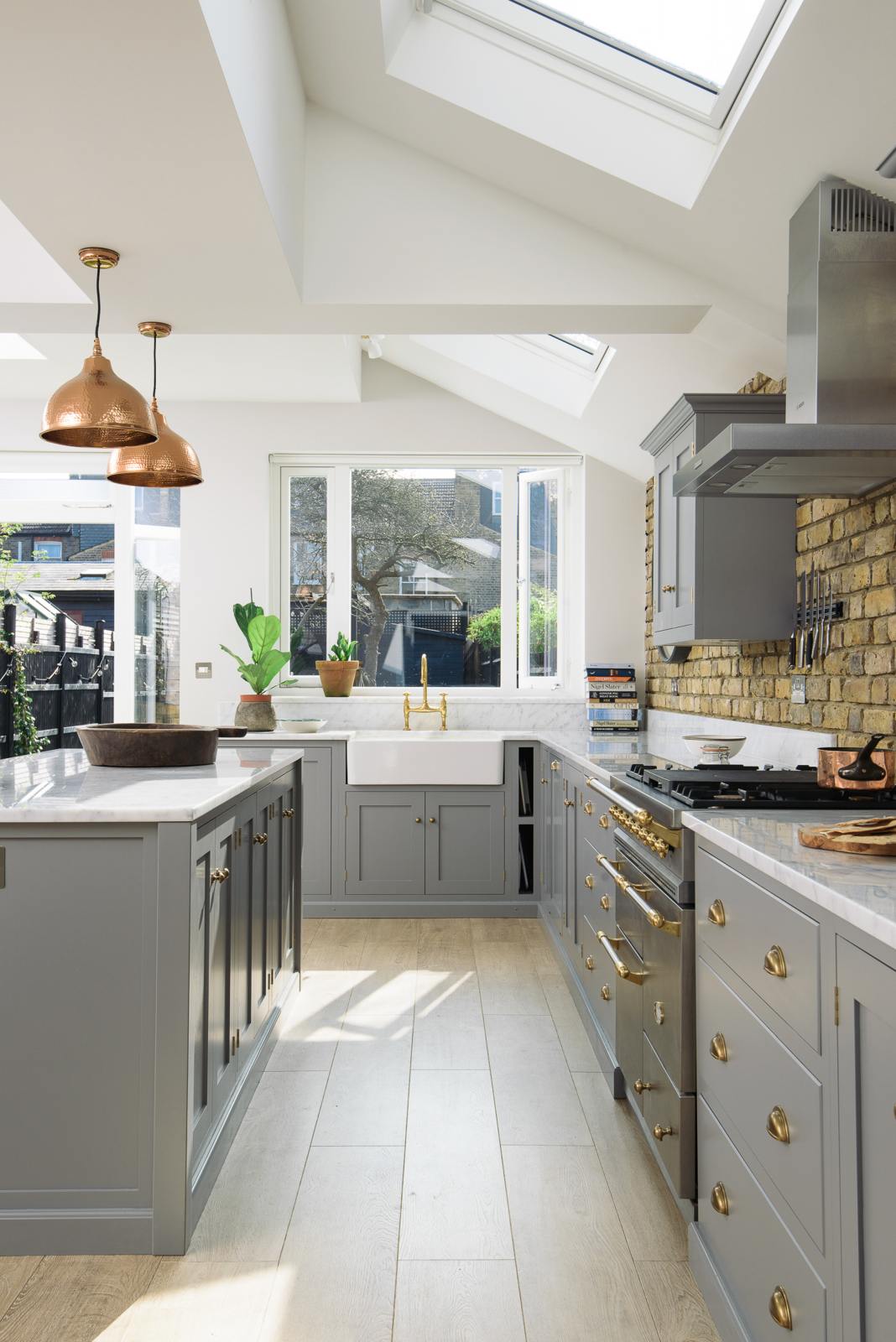Nestled between a soft industrial grey L-shaped sink and cooker run, which features a Carrara marble worktop, wall cupboards, and a kitchen island, is a single Belfast sink with an aged brass Ionian tap. Adorned with a weathered brick wall installation, copper pendant lights, simple ceramics, and colourful books, it creates a modern and functional kitchen for enthusiastic food connoisseurs.