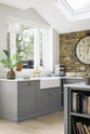 An oblique view of a soft industrial grey Shaker kitchen features an L-shaped sink and cooker run with a Carrara marble worktop, and a kitchen island. Adorned with a large pocket watch clock, a weathered brick wall installation, copper pendant lights, simple ceramics, and colourful books, it creates a modern and functional kitchen for enthusiastic food connoisseurs.