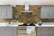 A soft industrial grey Shaker kitchen features an L-shaped sink and cooker run with a Carrara marble worktop. Adorned with a steel canopy, a weathered brick wall installation, a cream gas cooker, and a stone pestle and mortar, it creates a modern and functional kitchen for enthusiastic food connoisseurs.