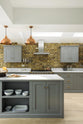 A soft industrial grey Shaker kitchen features an L-shaped sink and cooker run with a Carrara marble worktop, a kitchen island, wall cupboards, and a large silver refrigerator. Adorned with a steel canopy, a weathered brick wall installation, a cream gas cooker, and a stone pestle and mortar, it creates a modern and functional kitchen for enthusiastic food connoisseurs.
