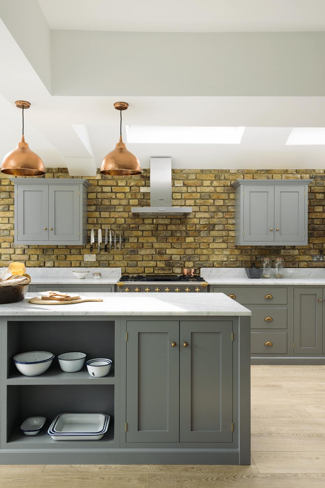 A soft industrial grey Shaker kitchen features an L-shaped sink and cooker run with a Carrara marble worktop, a kitchen island, wall cupboards, and a large silver refrigerator. Adorned with a steel canopy, a weathered brick wall installation, a cream gas cooker, and a stone pestle and mortar, it creates a modern and functional kitchen for enthusiastic food connoisseurs.