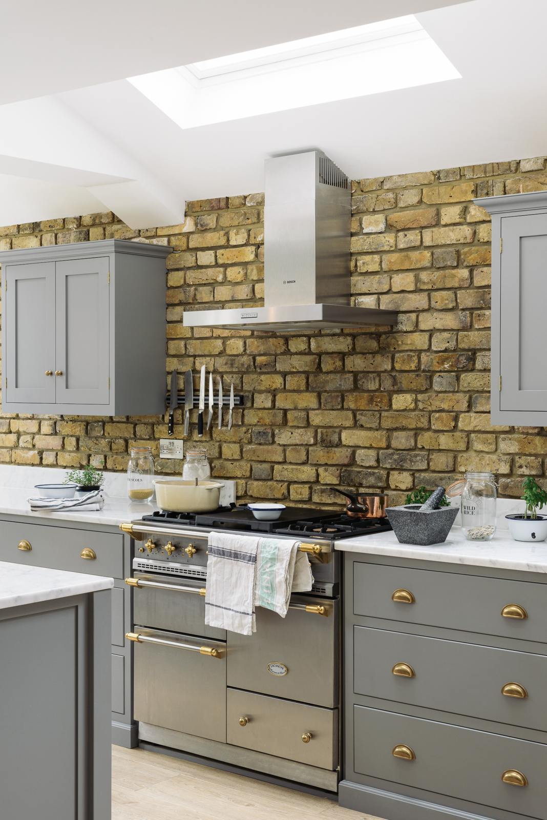 An oblique view of a soft industrial grey Shaker kitchen features an L-shaped sink and cooker run with a Carrara marble worktop, a kitchen island, wall cupboards, and a large silver refrigerator. Adorned with a steel canopy, a weathered brick wall installation, a cream gas cooker, and a stone pestle and mortar, it creates a modern and functional kitchen for enthusiastic food connoisseurs.