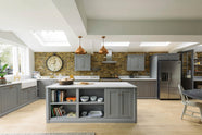 A soft industrial grey Shaker kitchen features an L-shaped sink and cooker run with a Carrara marble worktop, a kitchen island, wall cupboards, and a large silver refrigerator. Adorned with a steel canopy, a weathered brick wall installation, a cream gas cooker, and a stone pestle and mortar, it creates a modern and functional kitchen for enthusiastic food connoisseurs.