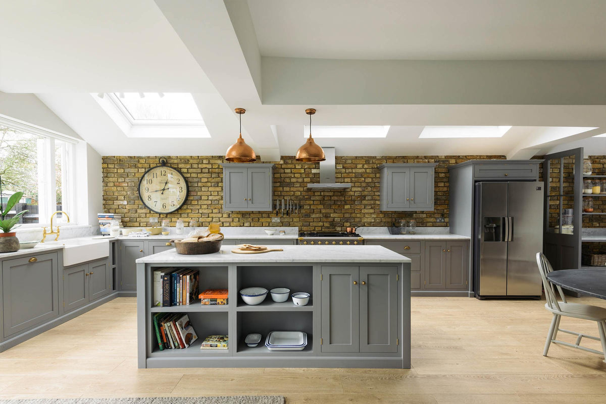 A soft industrial grey Shaker kitchen features an L-shaped sink and cooker run with a Carrara marble worktop, a kitchen island, wall cupboards, and a large silver refrigerator. Adorned with a steel canopy, a weathered brick wall installation, a cream gas cooker, and a stone pestle and mortar, it creates a modern and functional kitchen for enthusiastic food connoisseurs.