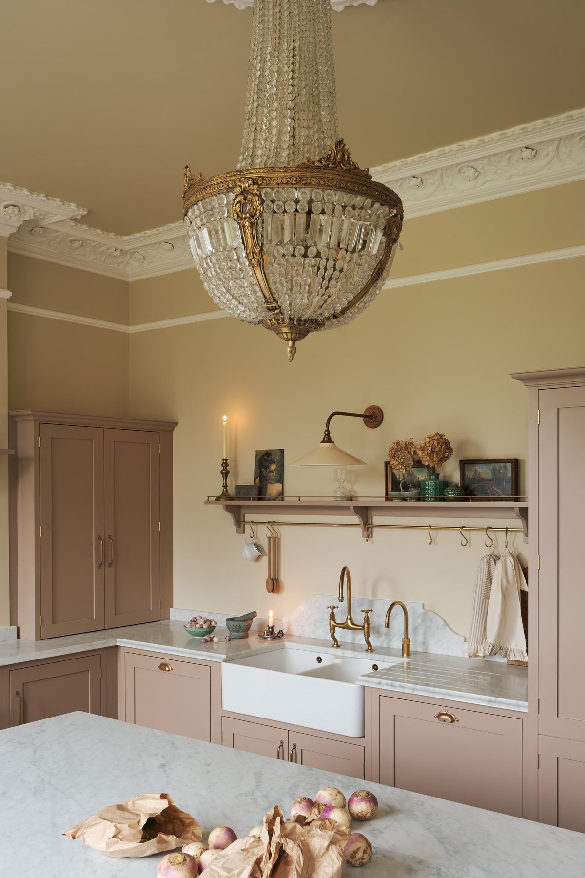 Looking past the large kitchen island of a romantic and peaceful Princelet pink Shaker kitchen is a tactile corner of a sink run, a countertop cupboard, shelf with brass railings, textured marble worktop, and a fridge and freezer cupboard. Beautifully paired with the subtle butter yellow painted walls, decorative cornice, a grand antique chandelier, and assorted vintage personal items, it creates a warm and inviting space.