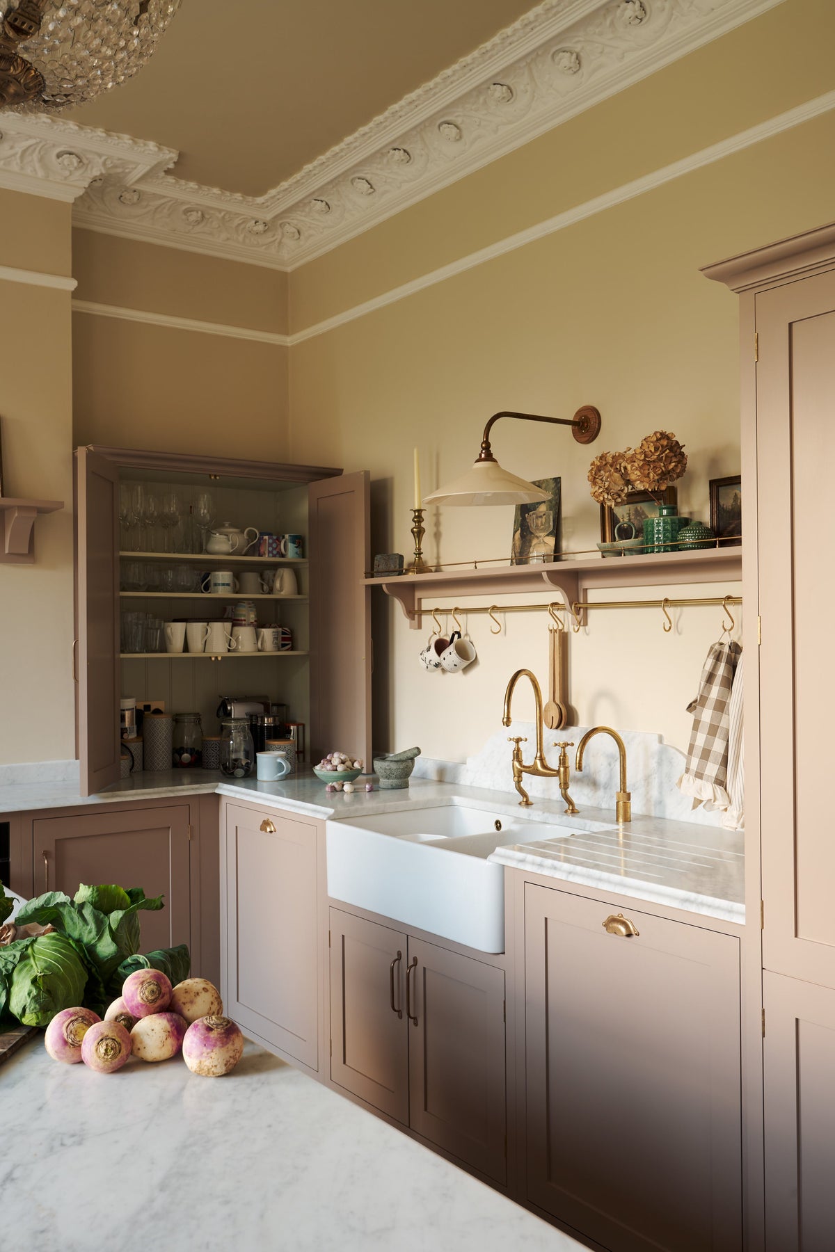 Past the large kitchen island of a romantic and peaceful Princelet pink Shaker kitchen is a tactile corner of a sink run, an open countertop cupboard, shelf with brass railings, textured marble worktop, and a fridge and freezer cupboard. Beautifully paired with the subtle butter yellow painted walls, decorative cornice, beautiful mugs and assorted vintage personal items, it creates a warm and inviting space.