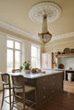 An oblique view of a romantic and peaceful Princelet pink Shaker kitchen features an L-shaped sink and cooker run with a marble worktop, matching countertop cupboards, a kitchen island with a breakfast nook and an overmantle above the chimney breast. Beautifully paired with the subtle butter yellow painted walls, decorative cornice and a grand antique chandelier, it creates a warm and inviting space.