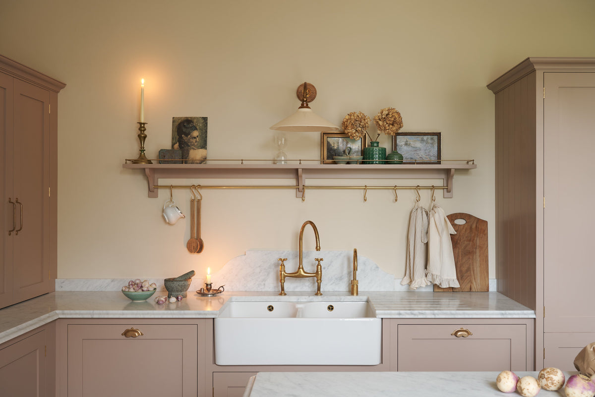 Past the large kitchen island of a romantic and peaceful Princelet pink Shaker kitchen is a tactile corner of a sink run, a countertop cupboard, shelf with brass railings, textured marble worktop, and a fridge and freezer cupboard. Beautifully paired with the subtle butter yellow painted walls, decorative cornice and assorted vintage personal items, it creates a warm and inviting space.