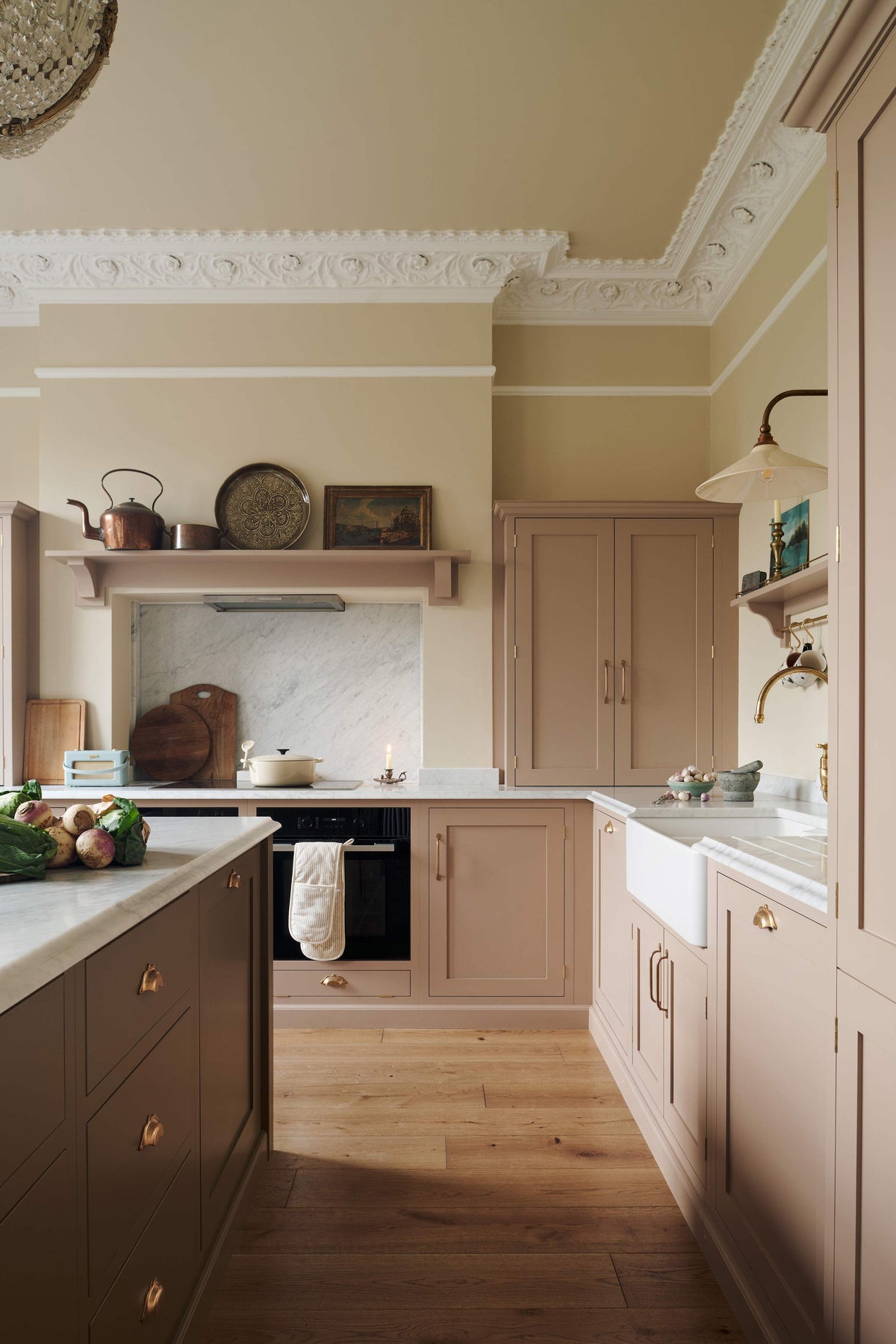 Past the large kitchen island of a romantic and peaceful Princelet pink Shaker kitchen is a quiet corner of a cooker run, a countertop cupboard, a marble worktop, an overmantle above the chimney breast, and a large white window sitting peacefully beside it. Beautifully paired with the subtle butter yellow painted walls, decorative cornice and assorted vintage personal items, it creates a warm and inviting space.
