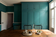 A grand, seafoam green Shaker kitchen featuring a bank of full-height pantry cupboards and a free-standing closed larder with Carrara marble worktop. Adorned with beautiful vintage furniture, a glass bowl of green figs, antique silverware, and a decanter, the kitchen reflects the refined tastes of its socialite and collector owners.