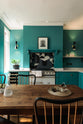A grand, seafoam green Shaker kitchen featuring an L-shaped cooker and sink run with a Carrara marble worktop, an overmantle canopy, and abstract splashback tiles. Adorned with beautiful vintage furniture, a luscious green house plant, a detailed block print, antique silverware, and a decanter, the kitchen reflects the refined tastes of its socialite and collector owners.