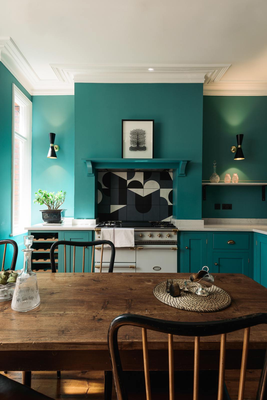 A grand, seafoam green Shaker kitchen featuring an L-shaped cooker and sink run with a Carrara marble worktop, an overmantle canopy, and abstract splashback tiles. Adorned with beautiful vintage furniture, a luscious green house plant, a detailed block print, antique silverware, and a decanter, the kitchen reflects the refined tastes of its socialite and collector owners.