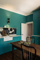 An angled view reveals a grand, seafoam green Shaker kitchen featuring an L-shaped cooker and sink run with a single Belfast sink, an aged brass Ionian tap, and a free-standing closed larder with Carrara marble worktop. Adorned with beautiful vintage furniture, a glass bowl of green figs, antique silverware, and a decanter, the kitchen reflects the refined tastes of its socialite and collector owners.