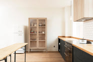 A simple and contemporary Sebastian Cox kitchen features a long sink and cooker run with a solid oak worktop, a single Belfast sink, an aged brass Ionian tap, a canopy, and a glazed freestanding cupboard. Adorned with bare pendant lights, an Oktoberfest bench and table, colourful ceramics, and a white refrigerator, this kitchen creates a cosy space with industrial details and a practical finish.