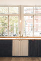 A simple and contemporary Sebastian Cox kitchen features a long sink run with a solid oak worktop, a single Belfast sink, and an aged brass Ionian tap. Adorned with bare pendant lights, a wooden cleaning brush, and a beautiful view of East London, this kitchen creates a cosy space with industrial details and a practical finish.