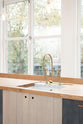 An oblique view of a simple and contemporary Sebastian Cox kitchen features a long sink run with a solid oak worktop, a single Belfast sink, and an aged brass Ionian tap. Adorned with bare pendant lights, a wooden cleaning brush, and a beautiful view of East London, this kitchen creates a cosy space with industrial details and a practical finish.