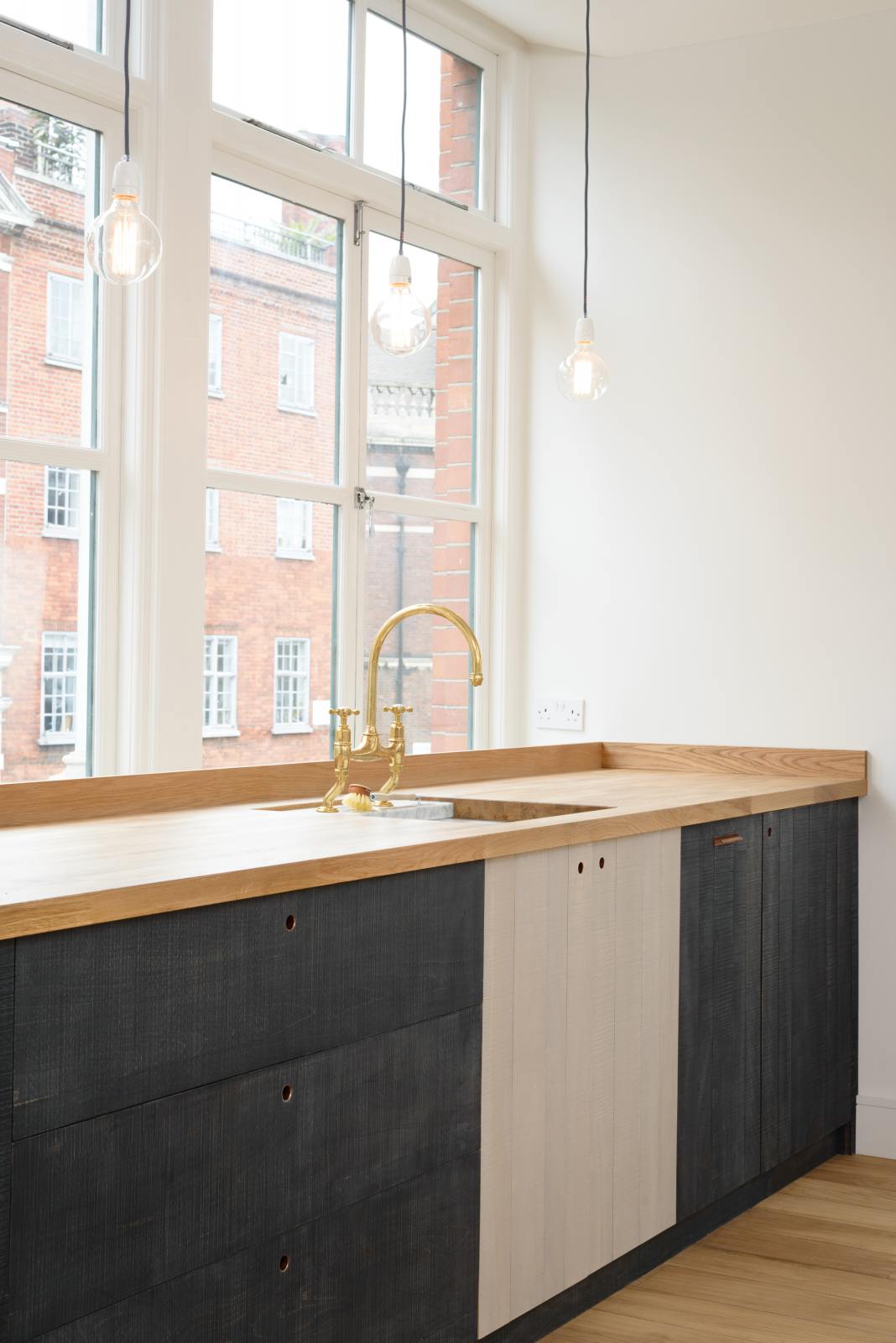 An oblique view of a simple and contemporary Sebastian Cox kitchen features a long sink, cooker run, a canopy, and a glazed freestanding cupboard. Adorned with bare pendant lights, an Oktoberfest bench and table, colourful ceramics, soft pillows, a metal beam structure, and a white refrigerator, this kitchen creates a cosy space with industrial details and a practical finish.