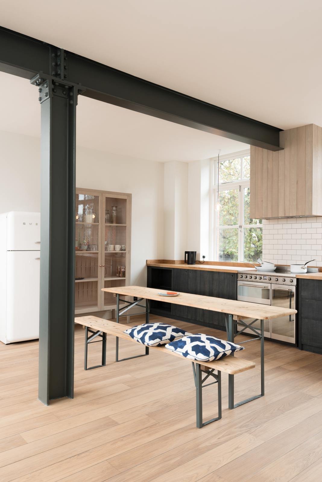 An oblique view of a simple and contemporary Sebastian Cox kitchen’s cooker run with a canopy, and a solid oak worktop. Adorned with bare pendant lights, a silver electrical hob, a black marble utensil holder, and a white refrigerator, this kitchen creates a cosy space with industrial details and a practical finish.