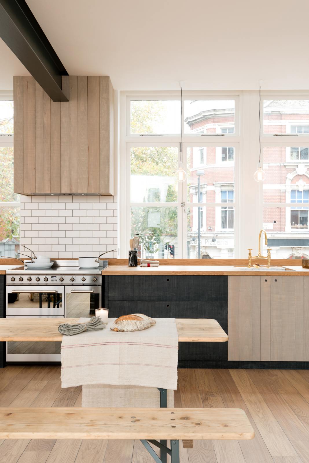 A simple and contemporary Sebastian Cox kitchen features a long sink and cooker run with a solid oak worktop, a single Belfast sink, an aged brass Ionian tap, a canopy, and a silver electric stove. Adorned with bare pendant lights, an Oktoberfest bench and table, white pans, a loaf of bread, and a natural linen table runner, this kitchen creates a cosy space with industrial details and a practical finish.