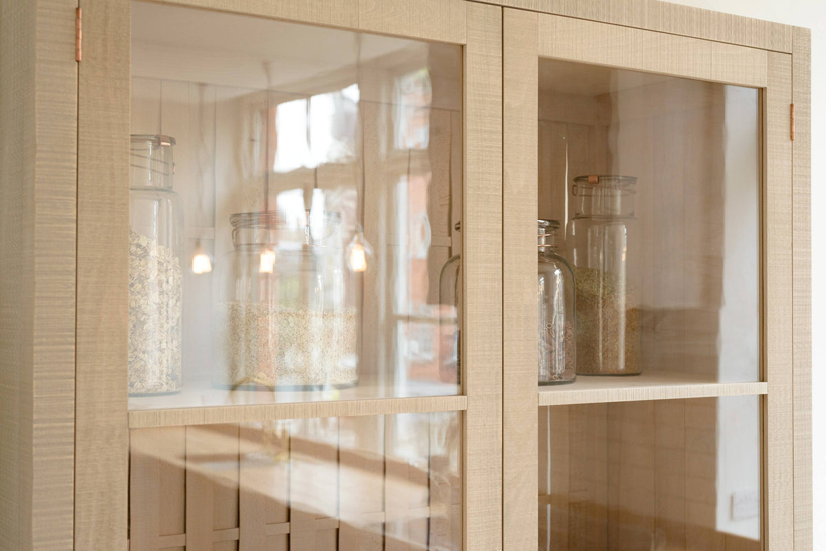 A close-up of a simple and contemporary Sebastian Cox kitchen’s glazed freestanding cupboard. Adorned with large glass containers of dried ingredients such as pasta. 
