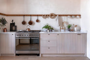 A warm and natural Sebastian Cox kitchen features a sink and cooker run with a Carrara marble worktop, a single Belfast sink, an aged brass Ionian tap, and a long oak peg rail. Beautifully decorated with artisan ceramics, copper pans, delicate potted plants, and blackened chopping boards, this kitchen creates a calm and delicate atmosphere that celebrates the use of natural materials.