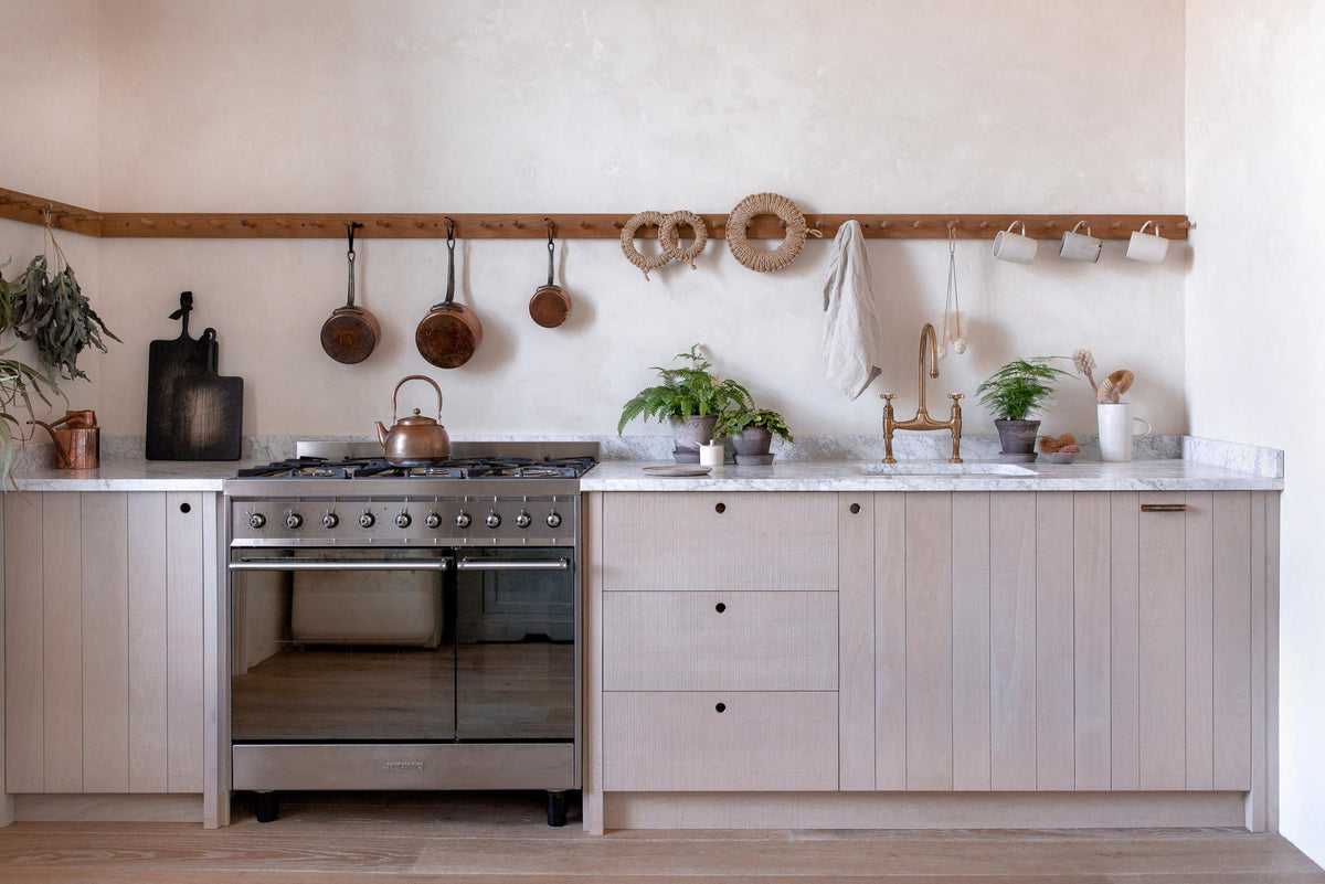 A warm and natural Sebastian Cox kitchen features a sink and cooker run with a Carrara marble worktop, a single Belfast sink, an aged brass Ionian tap, and a long oak peg rail. Beautifully decorated with artisan ceramics, copper pans, delicate potted plants, and blackened chopping boards, this kitchen creates a calm and delicate atmosphere that celebrates the use of natural materials.
