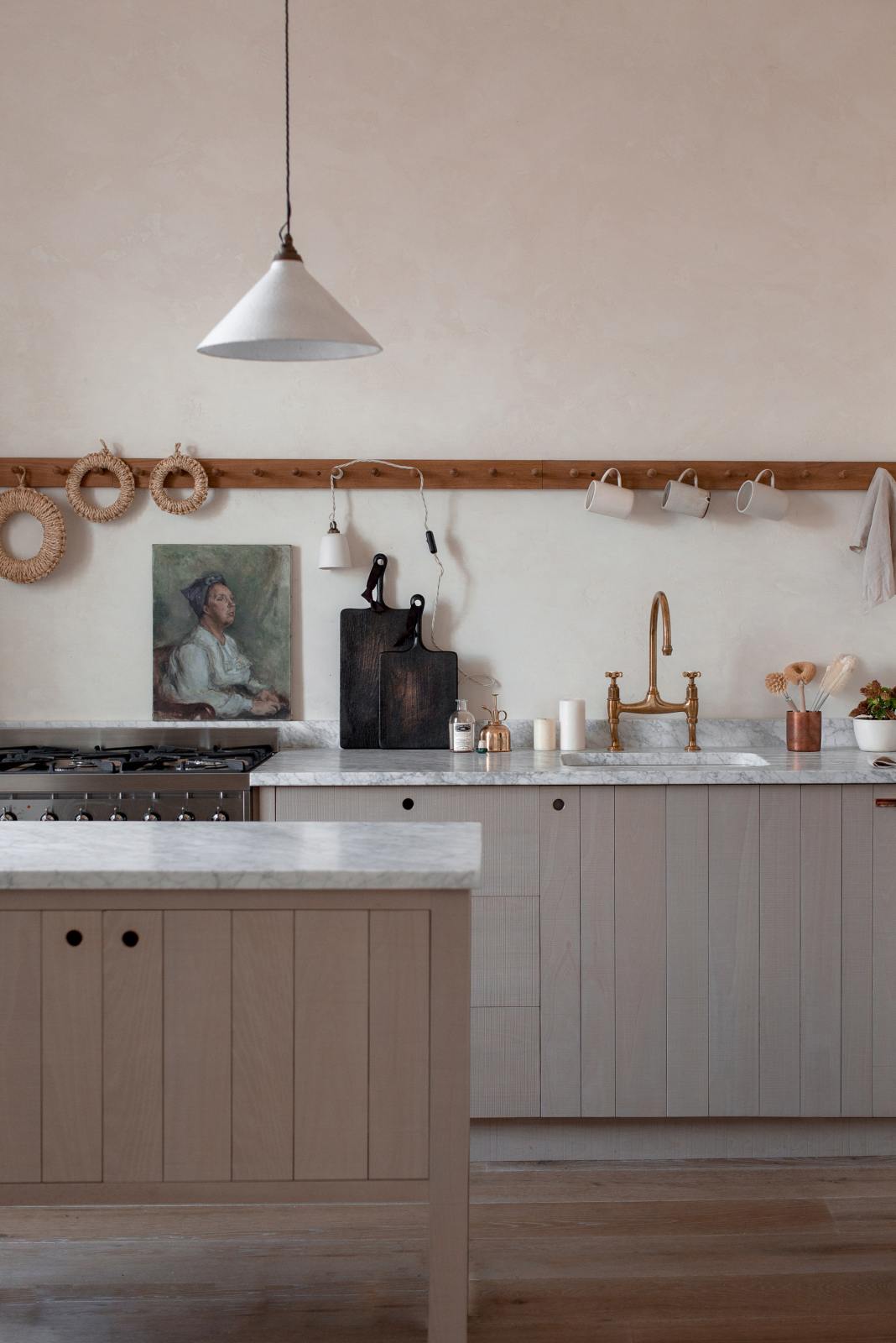A close-up of a warm and natural Sebastian Cox kitchen features a sink and cooker run with a Carrara marble worktop, a single Belfast sink, an aged brass Ionian tap, a long oak peg rail, and a movable prep table. Beautifully decorated with artisan ceramics, a creamware rise-and-fall light, a plug-in porcelain beaker light, and blackened chopping boards, this kitchen creates a calm and delicate atmosphere that celebrates the use of natural materials. | Image courtesy of Nina Plummer from INGREDIENTS LDN