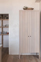 A quiet corner of a warm and natural Sebastian Cox kitchen features a movable prep table with a Carrara marble worktop, a freestanding cupboard, and a walk-in pantry. Beautifully decorated with artisan ceramics, drinking glasses, glass jars with dried ingredients, and a vintage bowl, this kitchen creates a calm and delicate atmosphere that celebrates the use of natural materials.