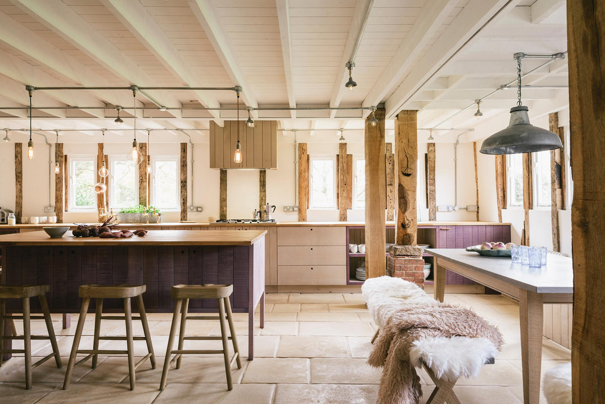 A soft and spacious Sebastian Cox barn kitchen features a long sink and cooker run with a solid oak worktop, two Belfast sinks, aged brass Mayan taps, a canopy, and a kitchen island with a breakfast nook. Beautifully decorated with lavender colour tints, bare pendant lights, vintage furniture, artisan ceramics, sweet potatoes, and sheep rugs, this kitchen creates an elegant and ethereal space.
