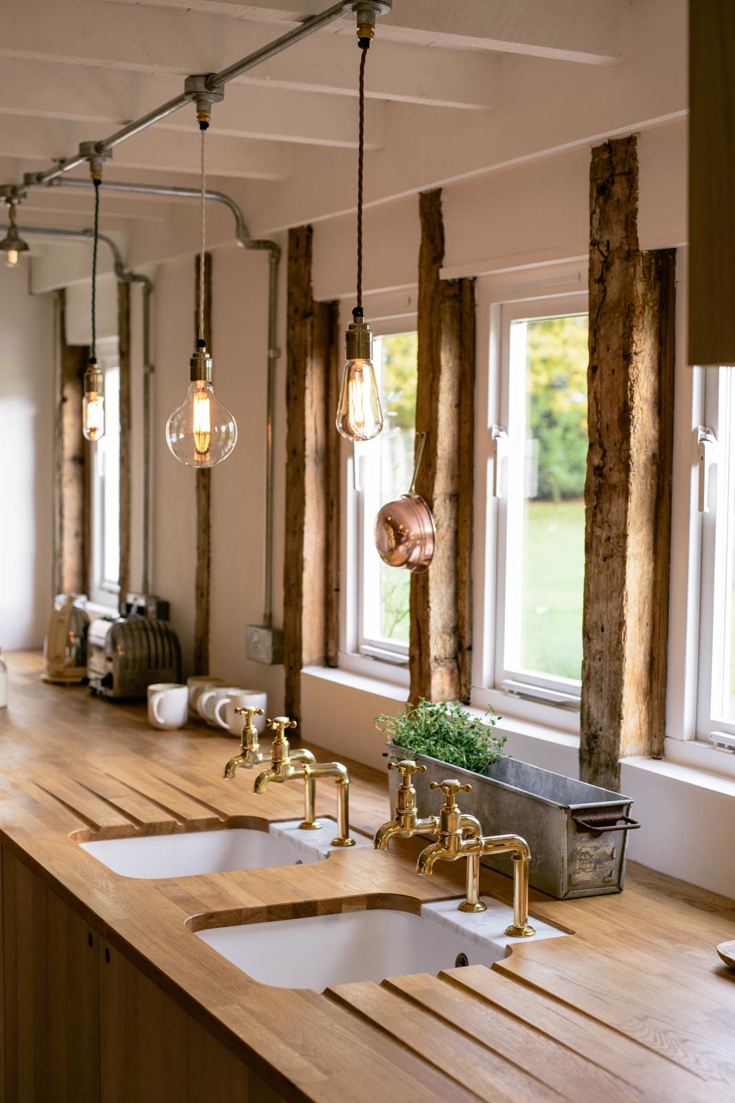 A close-up of a soft and spacious Sebastian Cox barn kitchen’s sink run features a solid oak worktop, two Belfast sinks, and aged brass Mayan taps. Beautifully decorated with lavender colour tints, bare pendant lights, artisan ceramics, and a copper strainer, this kitchen creates an elegant and ethereal space.