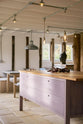 A closer view of a soft and spacious Sebastian Cox barn kitchen island with a breakfast nook. Beautifully decorated with lavender colour tints, bare pendant lights, vintage furniture, artisan ceramics, and organic beets, this kitchen creates an elegant and ethereal space.