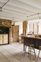 An angled view of a soft and spacious Sebastian Cox barn kitchen features a long sink and cooker run with a solid oak worktop, a kitchen island with a breakfast nook, and a muted yellow gas cooker. Beautifully decorated with lavender colour tints, bare pendant lights, artisan ceramics, wooden stools, and organic beets, this kitchen creates an elegant and ethereal space.