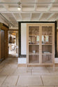 On the other side of a soft and spacious Sebastian Cox barn kitchen sits a glazed freestanding cupboard, beautifully decorated with artisan ceramics and drinking glasses. Past a small corridor is a quaint living room featuring rattan armchairs, a vintage wooden partition, and a large oak pantry cupboard.