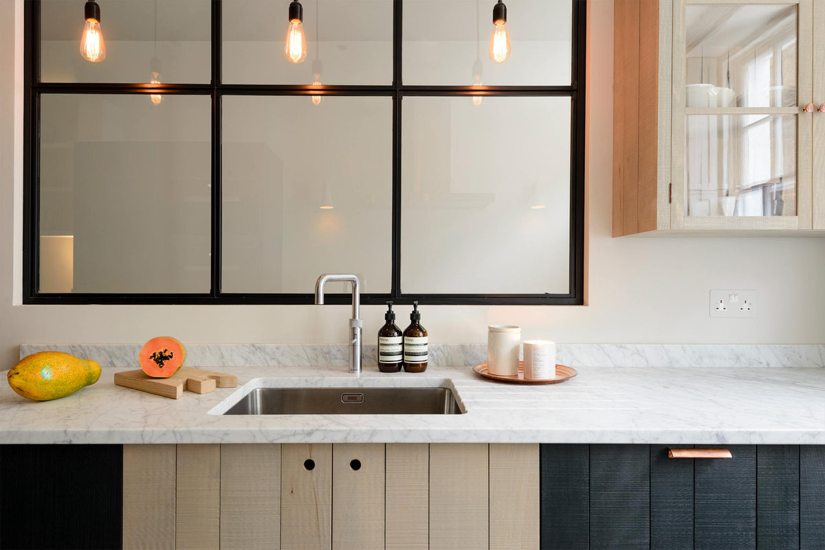 A sleek and modern Sebastian Cox galley kitchen features a sink run with Carrara marble worktop, a transom partition window, and a glazed wall cupboard. Adorned with simple ceramics, bare pendant lights, a bright orange papaya on a thick chopping board, and a candle, this kitchen creates an elegant and practical space.