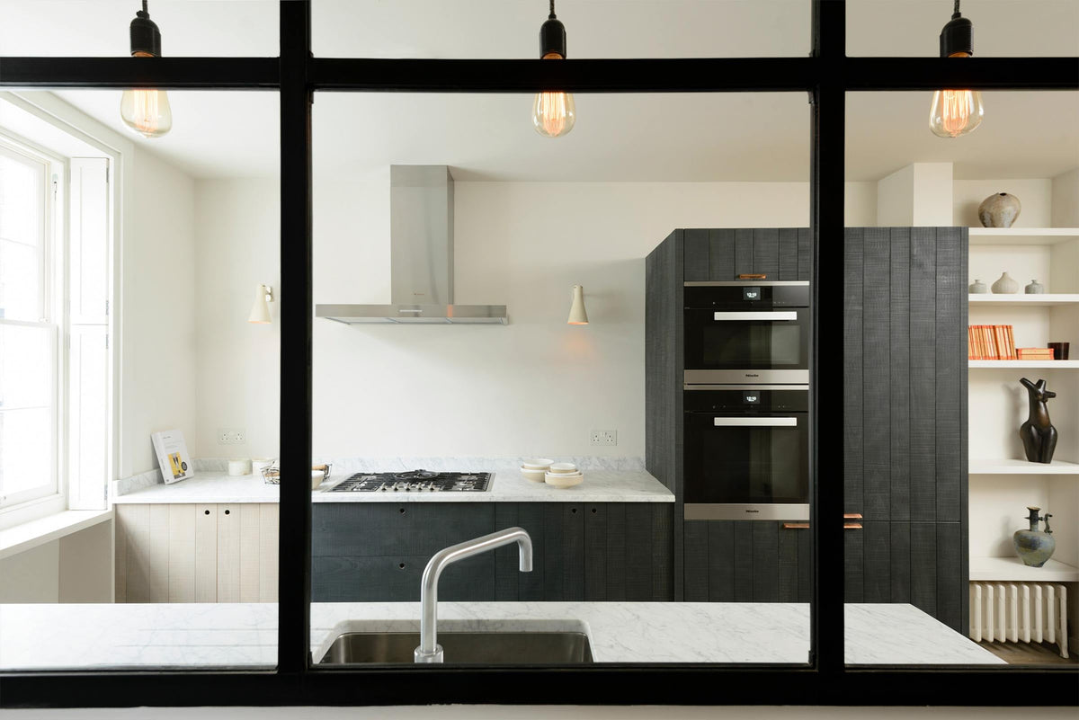 Past a black-framed transom partition window is a sleek and modern Sebastian Cox galley kitchen featuring a sink and cooker run with a Carrara marble worktop, an appliance cabinet, and a fridge/freezer cupboard. Adorned with simple ceramics, decorative ceramics, bare pendant lights, a metal canopy, and books, this kitchen creates an elegant and practical space.