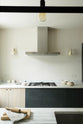 Past a black-framed transom partition window is a sleek and modern Sebastian Cox galley kitchen featuring a sink and cooker run with a Carrara marble worktop. Adorned with simple ceramics, beaker wall lights, a metal canopy, a chopping board, a rolling pin, and tea towels, this kitchen creates an elegant and practical space.