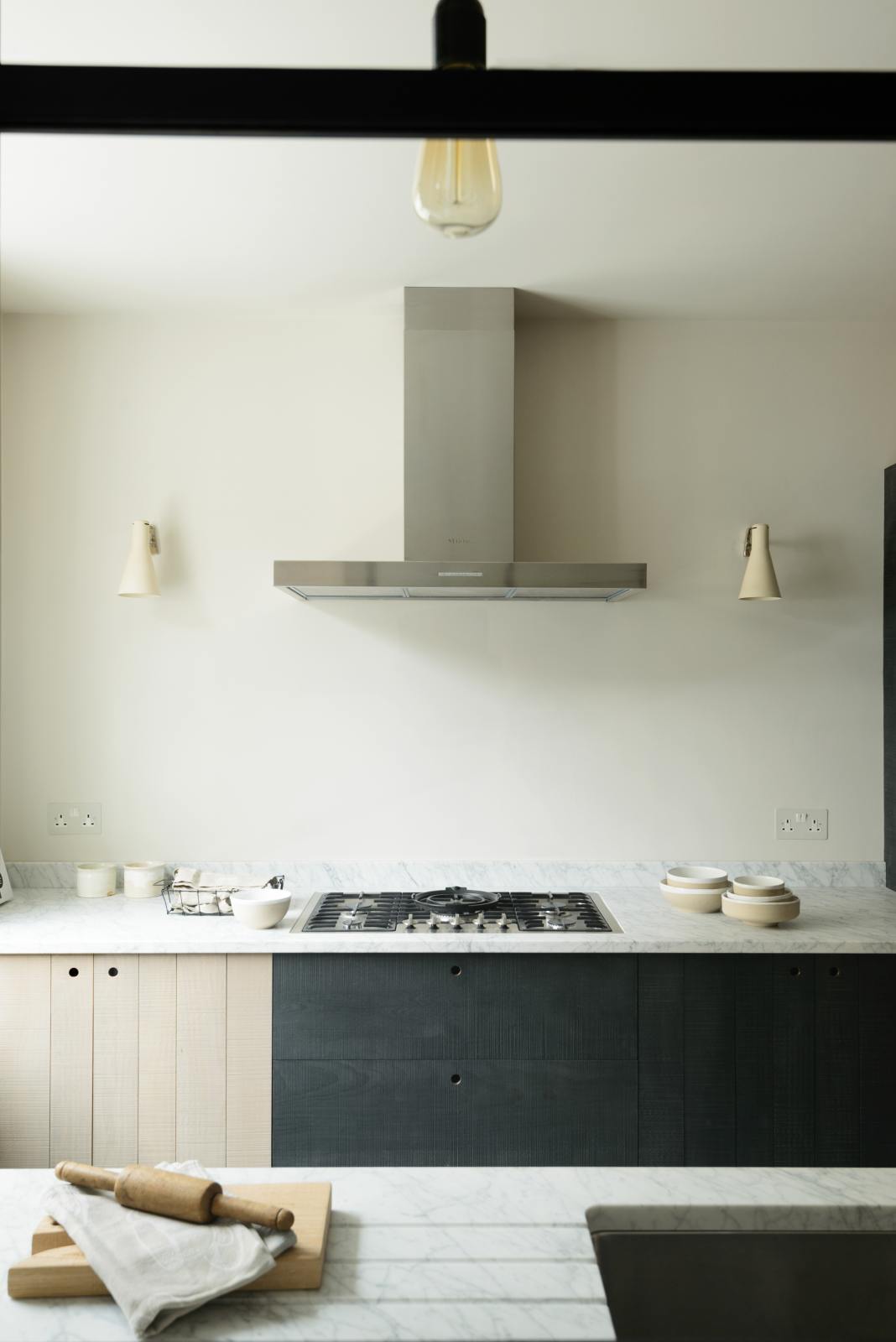 Past a black-framed transom partition window is a sleek and modern Sebastian Cox galley kitchen featuring a sink and cooker run with a Carrara marble worktop. Adorned with simple ceramics, beaker wall lights, a metal canopy, a chopping board, a rolling pin, and tea towels, this kitchen creates an elegant and practical space.