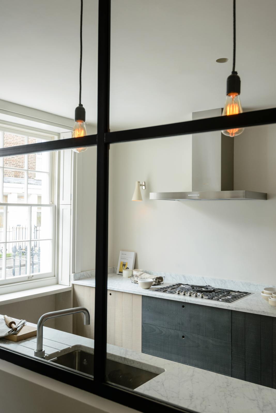 An oblique view past a black-framed transom partition window is a sleek and modern Sebastian Cox galley kitchen featuring a sink and cooker run with a Carrara marble worktop. Adorned with simple ceramics, beaker wall lights, a metal canopy, a chopping board, a rolling pin, and tea towels, this kitchen creates an elegant and practical space.