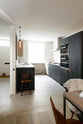 An oblique view of a sleek and modern Sebastian Cox galley kitchen features a sink and cooker run with Carrara marble worktop, a transom partition window, a glazed wall cupboard, an appliance cabinet, and a fridge/freezer cupboard. Adorned with simple ceramics, bare pendant lights, a wooden dining table and chairs, and a fluffy wool blanket, this kitchen creates an elegant and practical space.