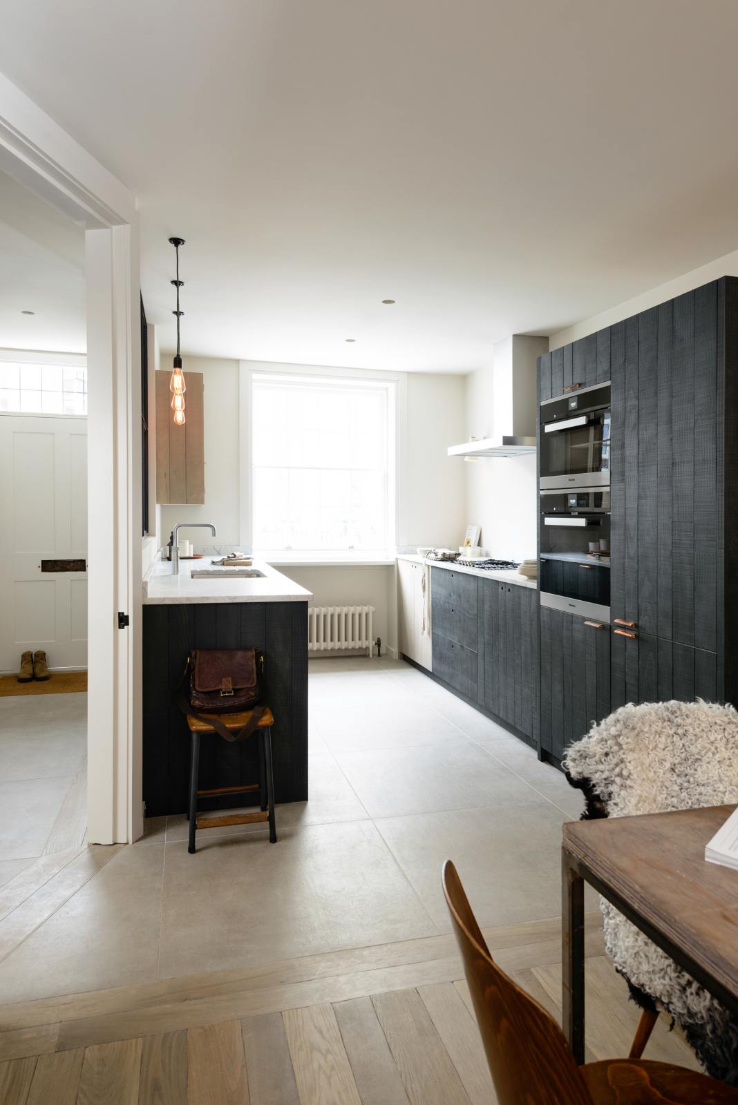 An oblique view of a sleek and modern Sebastian Cox galley kitchen features a sink and cooker run with Carrara marble worktop, a transom partition window, a glazed wall cupboard, an appliance cabinet, and a fridge/freezer cupboard. Adorned with simple ceramics, bare pendant lights, a wooden dining table and chairs, and a fluffy wool blanket, this kitchen creates an elegant and practical space.