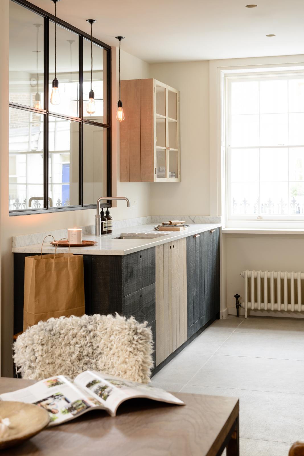 An oblique view of a sleek and modern Sebastian Cox galley kitchen features a sink run with Carrara marble worktop, a transom partition window, and a glazed wall cupboard. Adorned with simple ceramics, bare pendant lights, a wooden dining table and chairs, and a fluffy wool blanket, this kitchen creates an elegant and practical space.