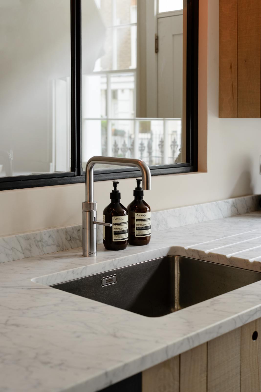 A close-up of a sleek and modern Sebastian Cox galley kitchen’s sink run features a transom window, a stainless steel sink, a silver Quooker tap, hand-wash bottles, and a Carrara marble worktop.