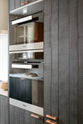 A close-up of a sleek and modern Sebastian Cox galley kitchen features an appliance cabinet and a fridge/freezer cupboard. Adorned with warm copper bar handles, these elements stand out against the inky black-blue tint of the natural wood grains of the exquisitely bespoke cupboard.