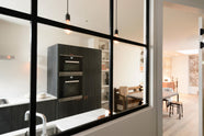 Past a black-framed transom partition window is a sleek and modern Sebastian Cox galley kitchen featuring a sink and cooker run with a Carrara marble worktop, an appliance cabinet, a fridge/freezer cupboard, and a dining space. Adorned with simple ceramics, decorative ceramics, books, bare pendant lights, mismatched vintage dining furniture, and a fluffy wool blanket, this kitchen creates an elegant and practical space.