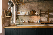 A calm and simple Sebastian Cox kitchen features a long sink run with two sets of copper sinks and aged brass Mayan taps, a copper worktop, a shelf, and a prep table with a solid oak worktop. Adorned with vintage items, artisan ceramics, wooden chopping boards, framed illustrations, tea towels, and wildflowers in large glass containers, this kitchen creates a chic New York industrial space that celebrates a love of natural materials.